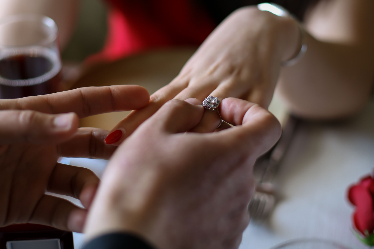 A man sliding a diamond engagement ring onto a woman’s finger at the dinner table, a glass of wine and a red rose visible in the background.