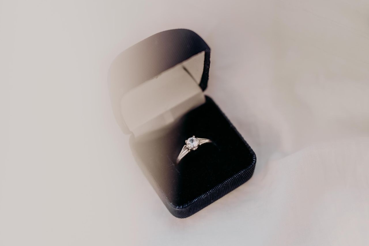 A beautiful engagement ring inside of a black velvet ring case, on top of a white table.