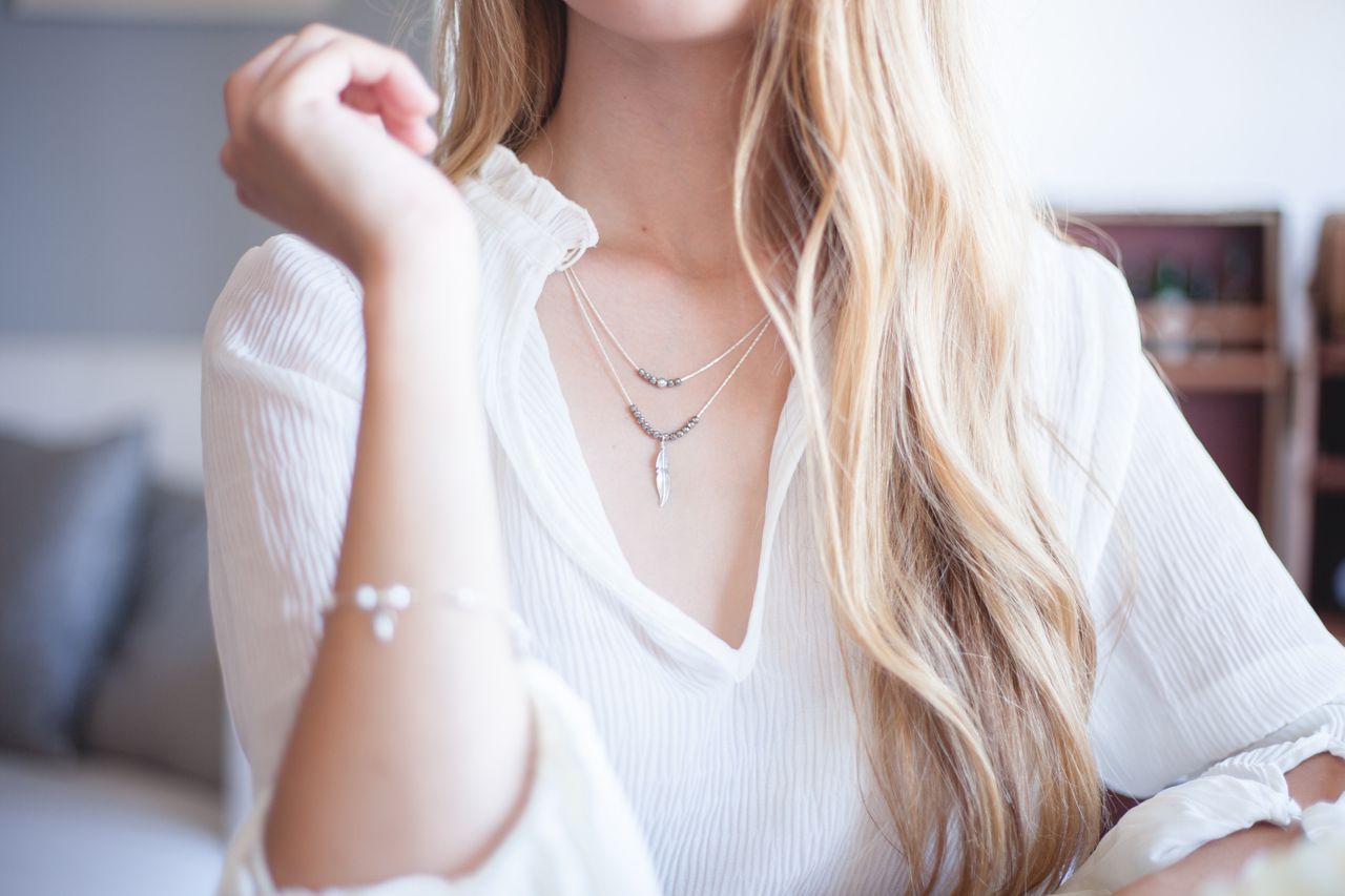 A woman in a white blouse sitting in a lobby, her stacked necklaces shining in the sun and her wrist adorned with a diamond bracelet.