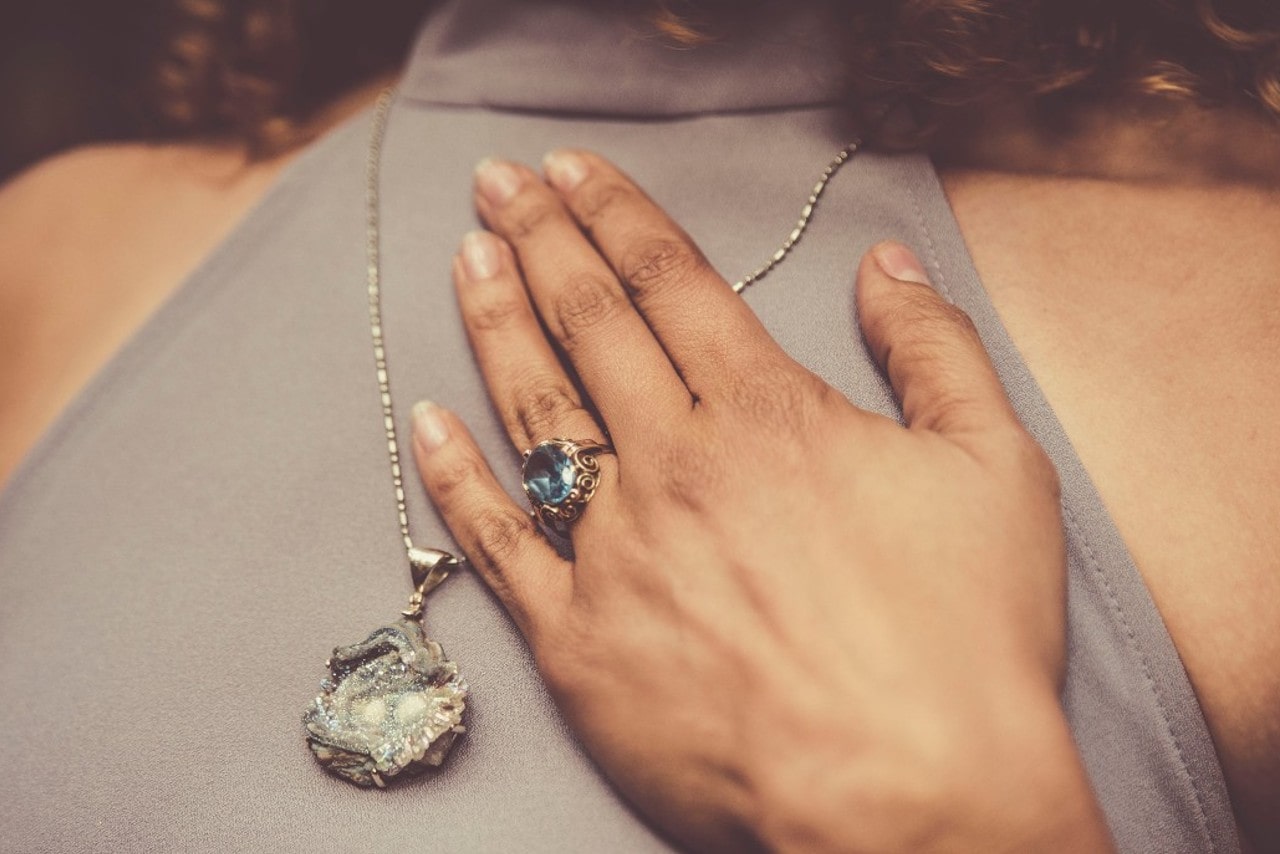 A woman wresting her hand on her chest overtop of a large pendant, a big blue stone on her ring finger.