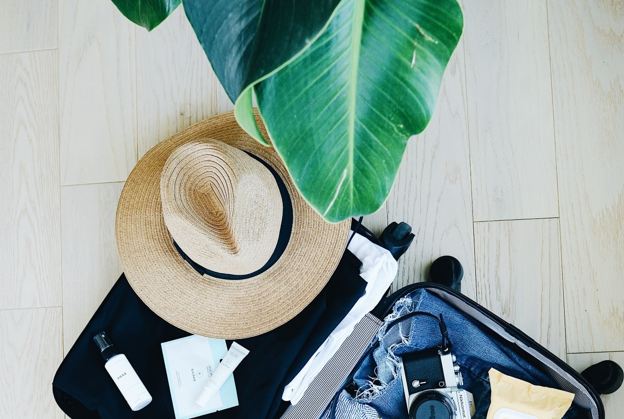 An open suitcase full of clothes, creams, camera and hat on top of the bright wooden floor