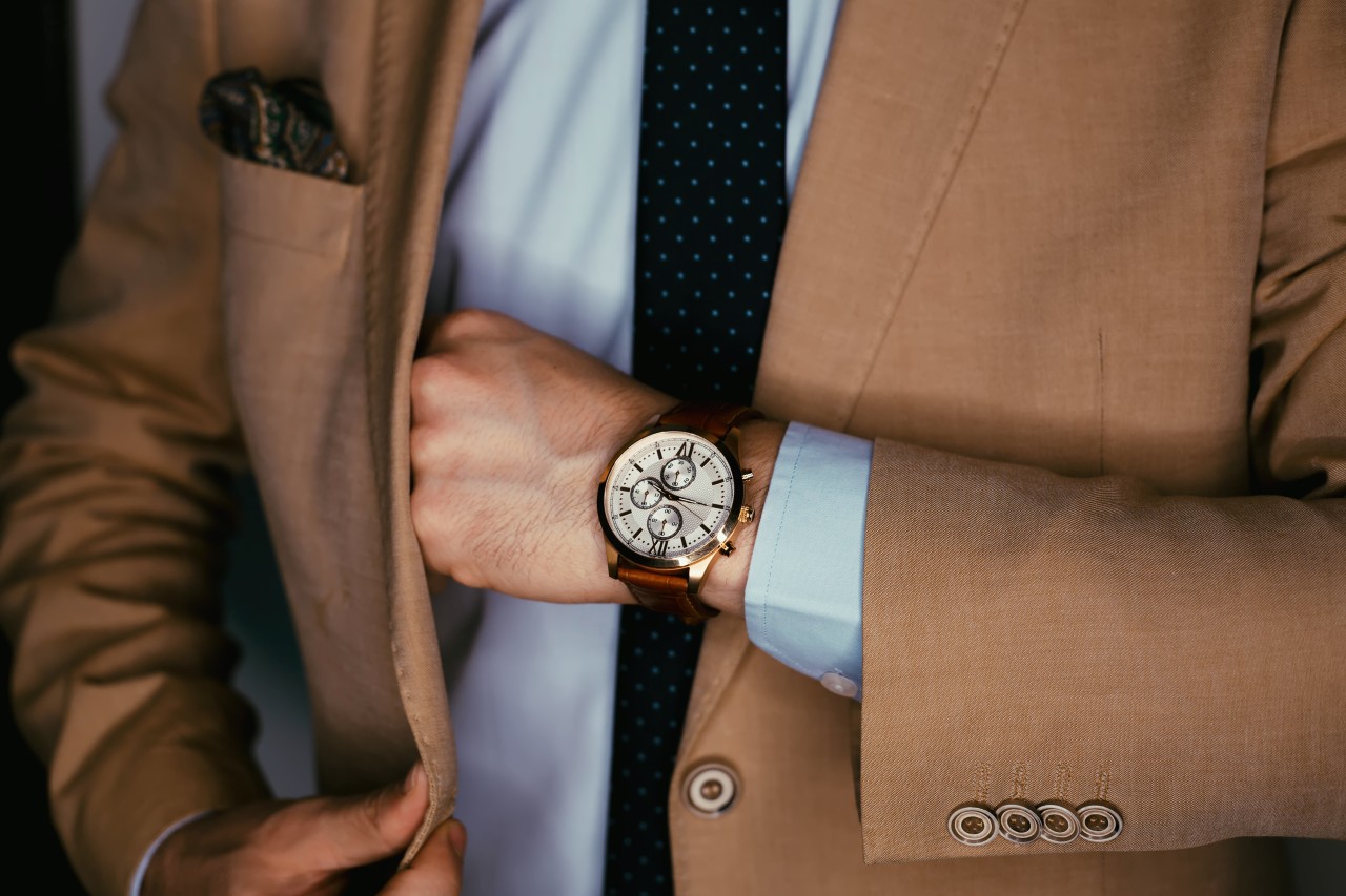 A man in a beige suit takes something from his inside pocket showcasing a leather watch with 3 subdials.