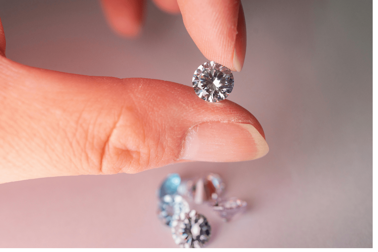 A close up of a loose diamonds held between two fingers, while other diamonds are scattered on a white surface