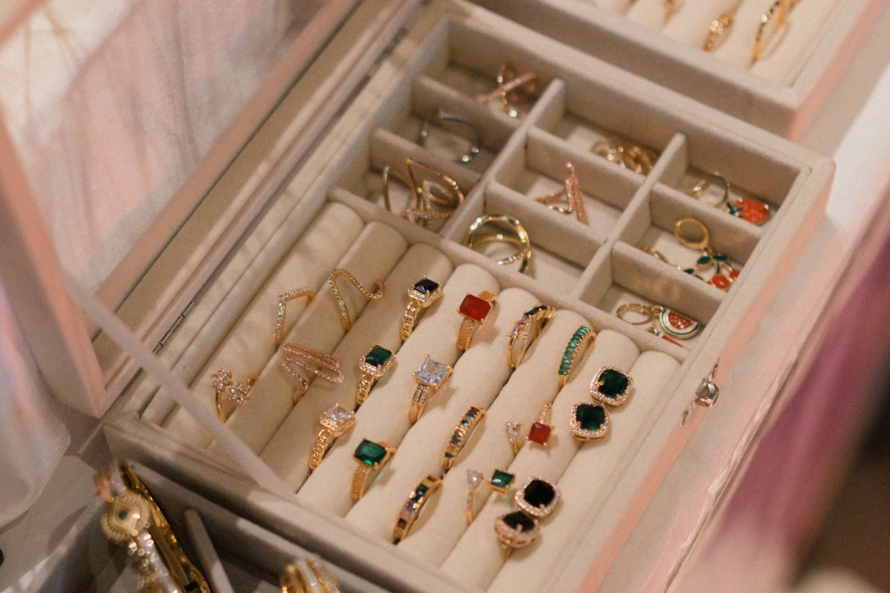 A jewelry box displays an arranged gold rings set with colorful gemstones, including emeralds, rubies, and clear stones, organized in soft velvet compartments.