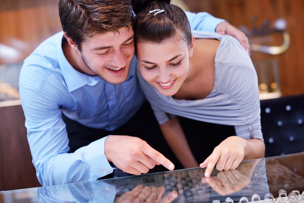 A happy couple leans over a glass display case, smiling and pointing excitedly at engagement rings inside a jewelry store.