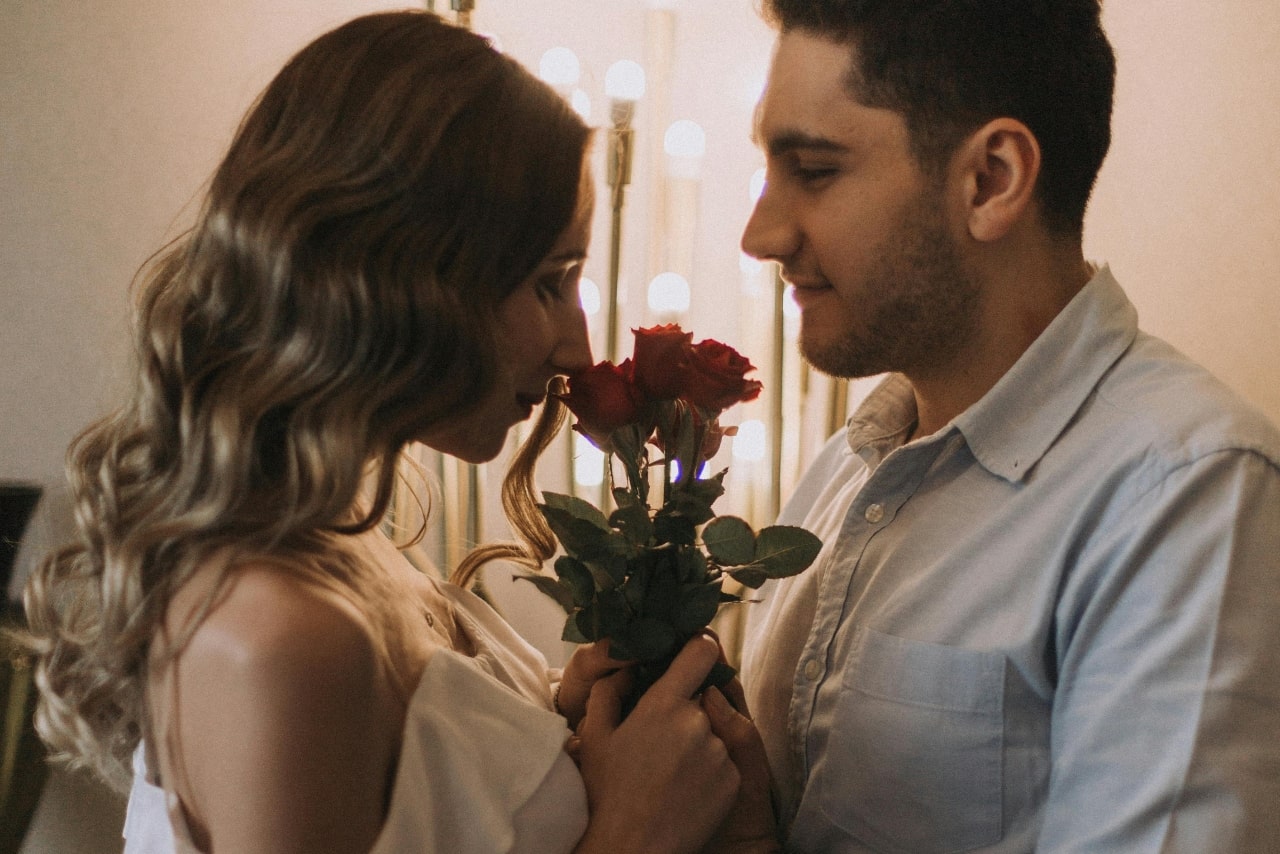 A couple standing closely to one another, the woman smelling red roses as the man looks lovingly at her.