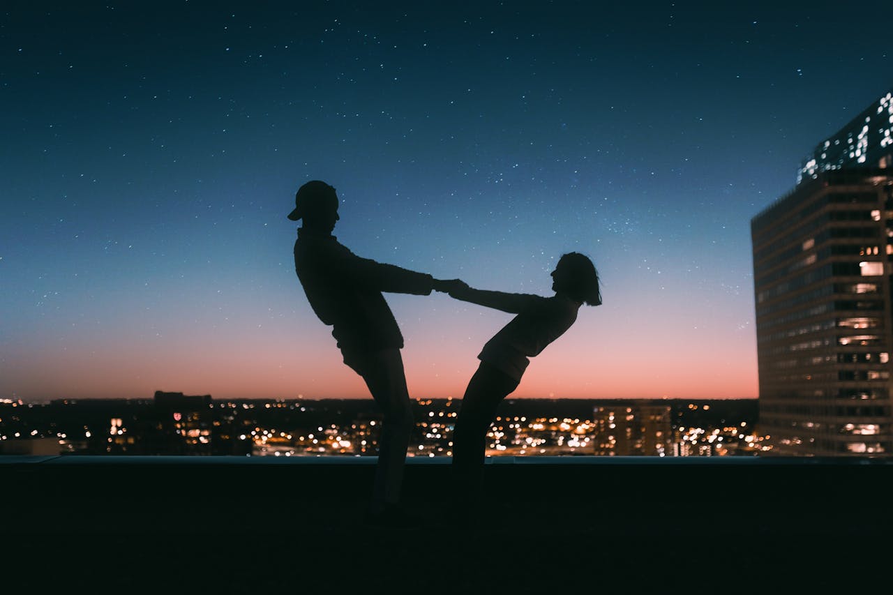 A backlit image of a couple dancing on a rooftop, the city skyline lit up behind them as the sun goes down.