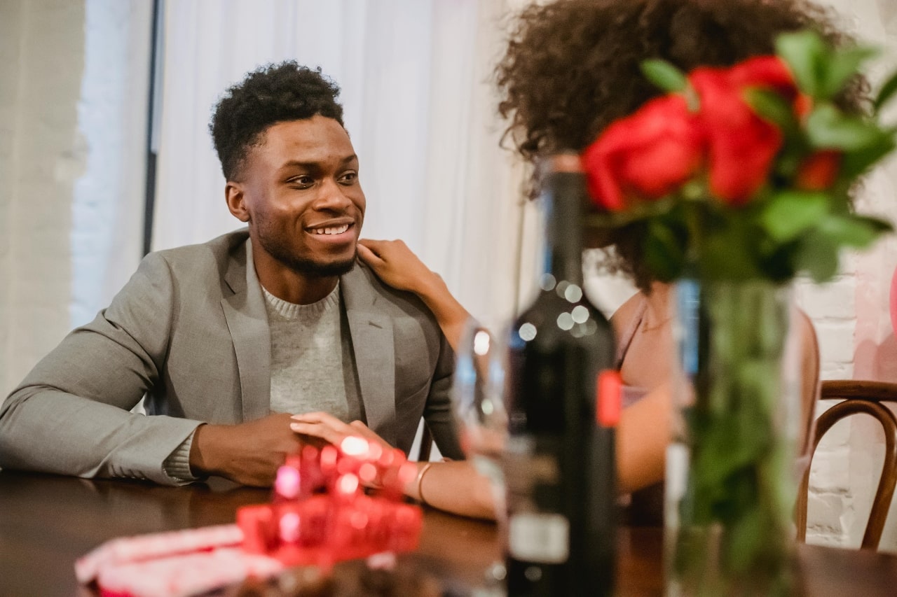A couple sitting at a table together with gifts, roses, and a bottle of wine.