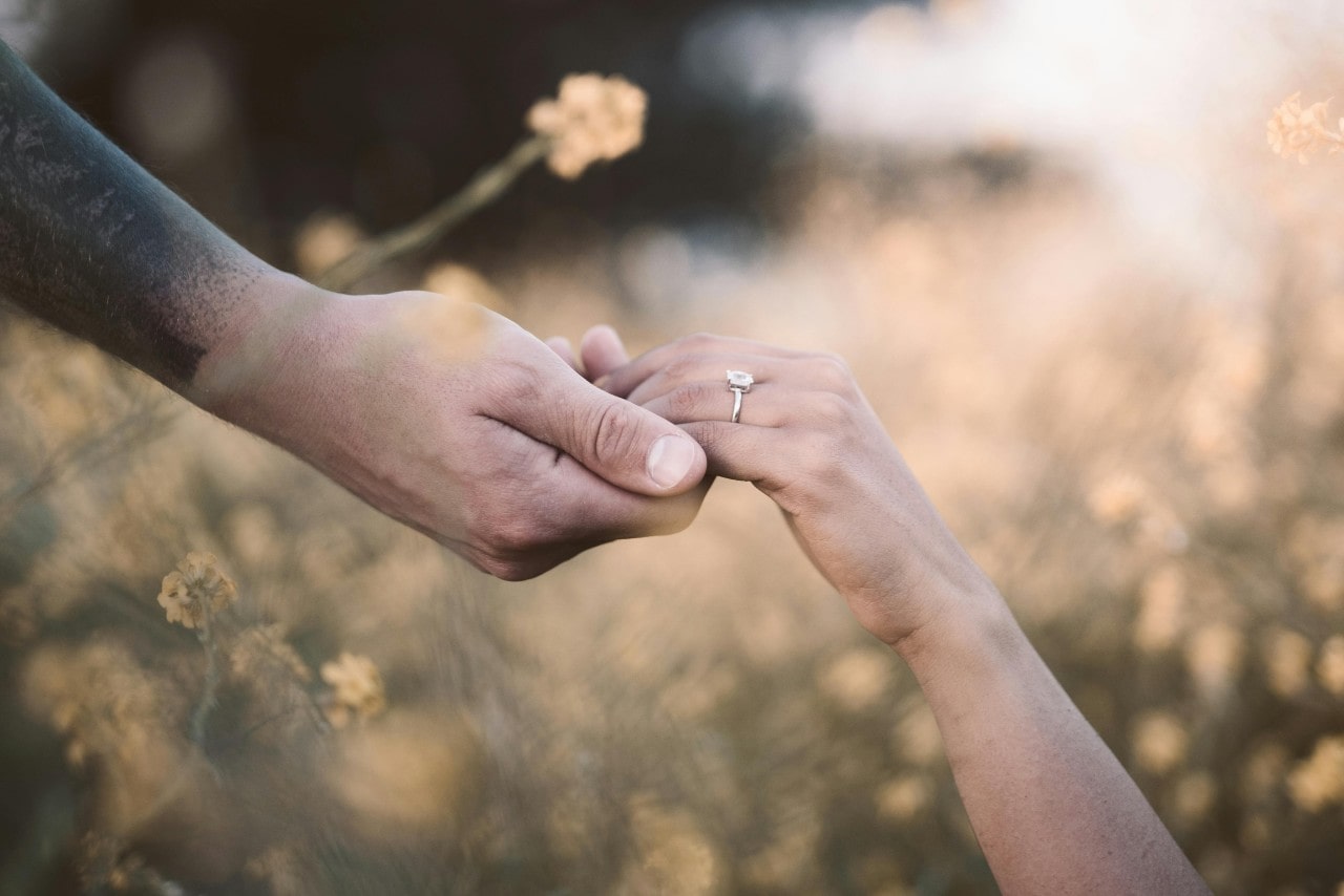 A close-up of a man holding a woman’s hand in a field of yellow flowers, her engagement ring being showcased.