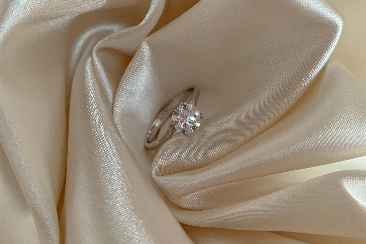 A close-up of a diamond solitaire engagement ring on a piece of shiny beige fabric.