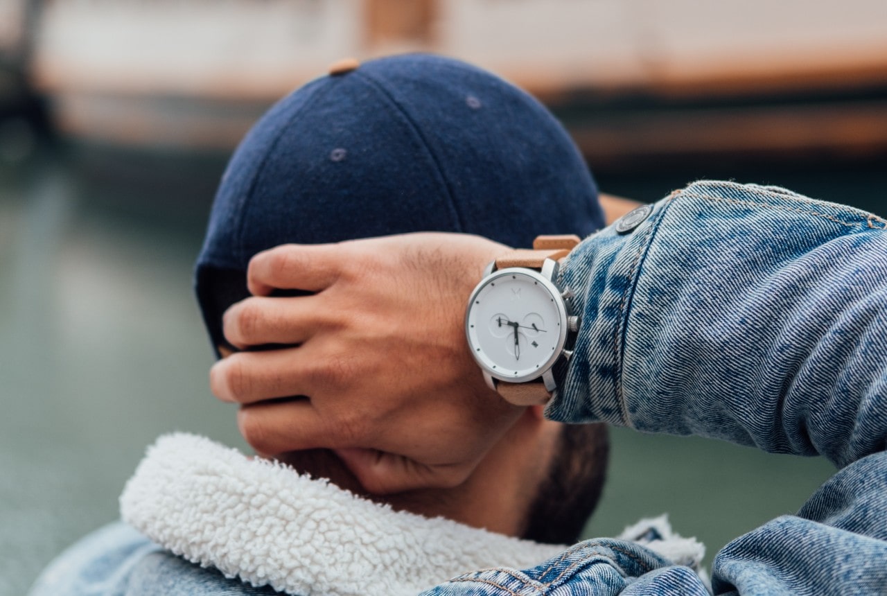A close-up of the back of a comfortably dressed man’s head. His hand, adorned with a luxury watch, touching the back of his cap.