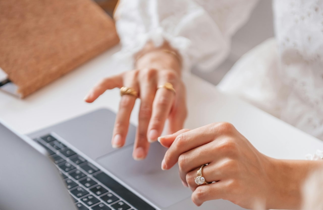 A close-up of a woman’s hands, adorned with gold rings and a large diamond engagement ring, typing on a laptop.