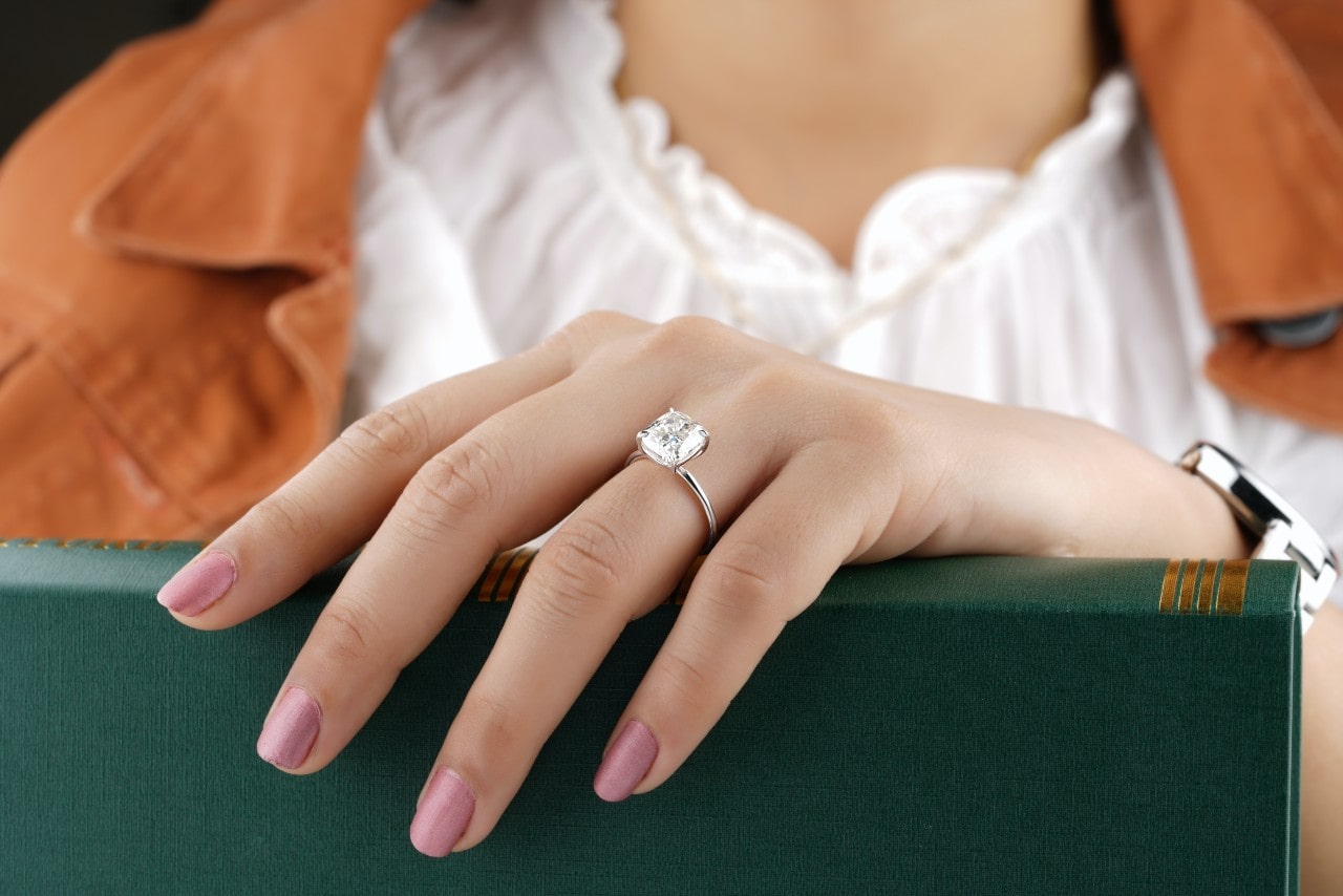 A close-up of a woman’s manicured hand resting on a green book, a solitaire diamond ring on her finger.