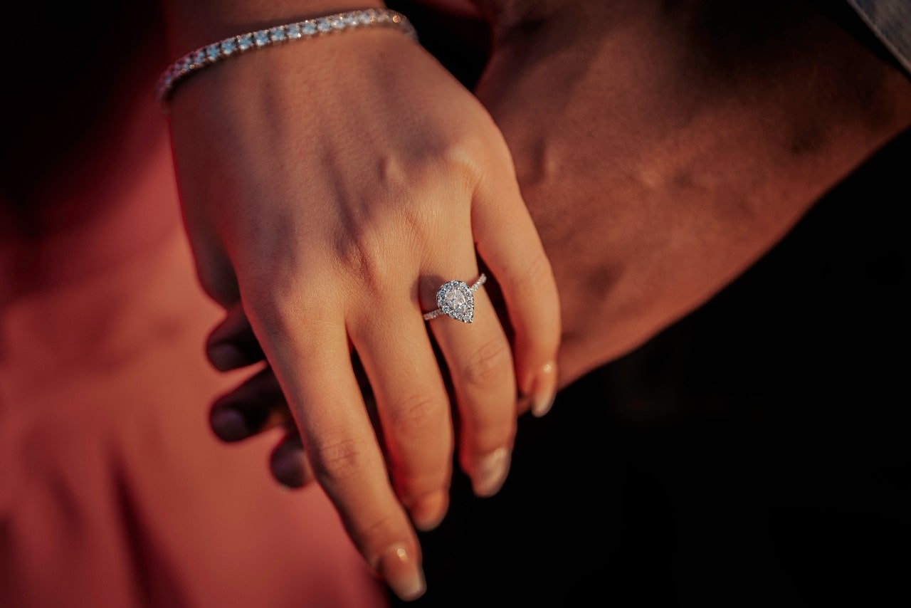 A close-up of a couple holding hands, the woman’s hand adorned with a halo engagement ring with a pear cut center stone.