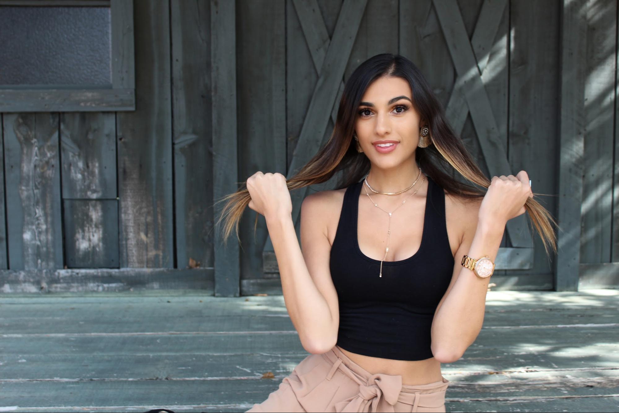 A smiling woman wearing fashion jewelry is posing in a black crop top and beige pants against a wooden backdrop.