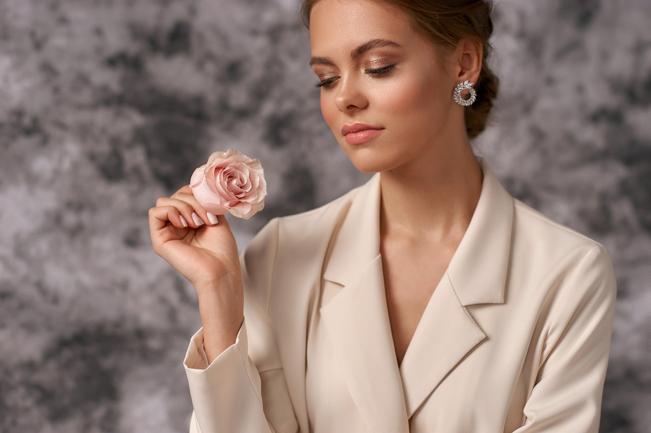 A woman in beige blaze holds a pink rose and showcases round white gold diamond earrings against a grey blurry background