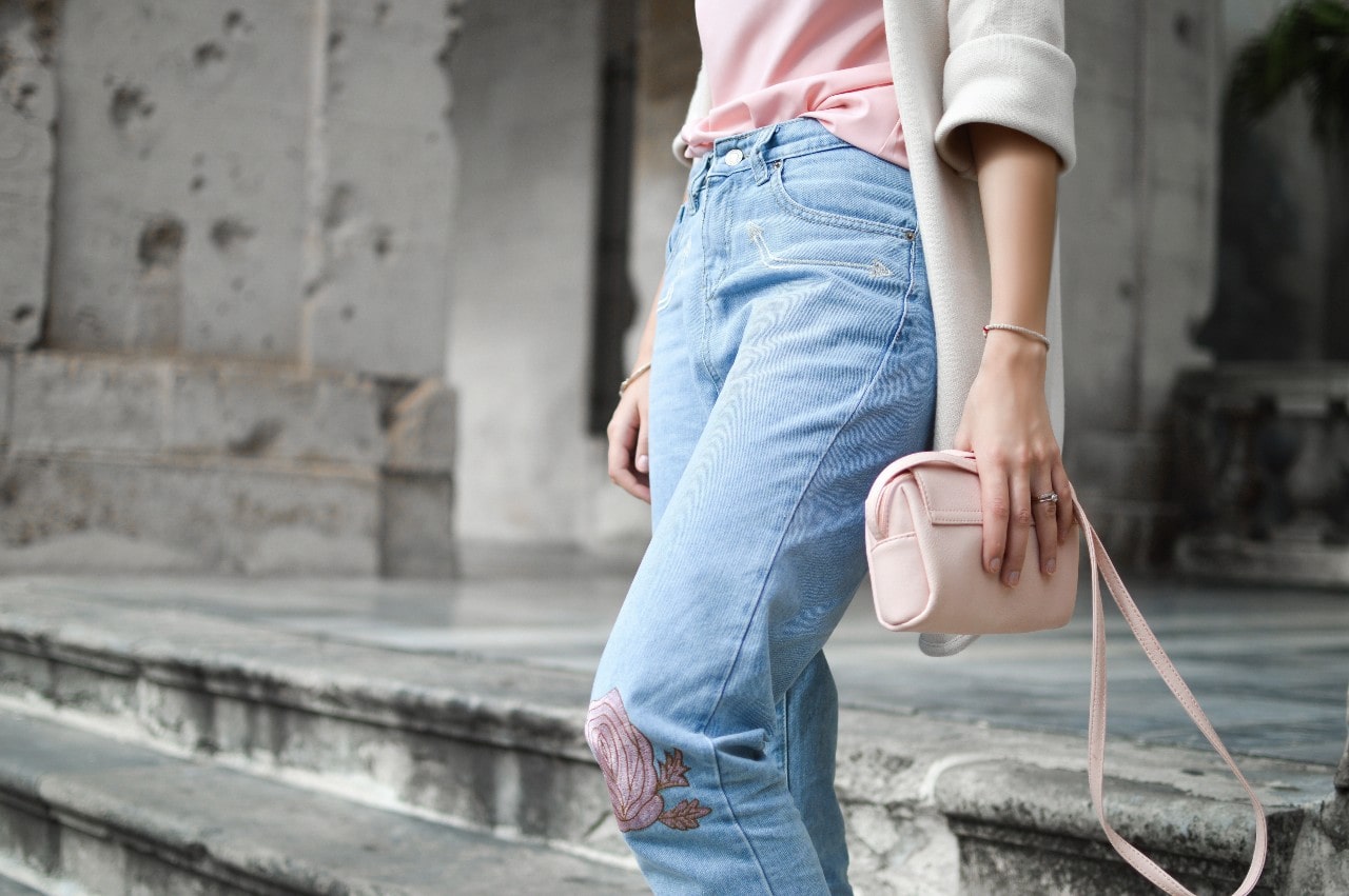 A woman in a pink shirt and blue jeans, holding a pink purse with a small tennis bracelet on her wrist, standing on stone steps.