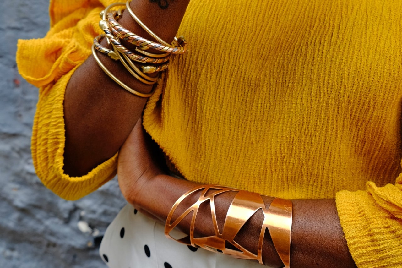 A close-up of a woman's arm adorned with multiple yellow gold bangles, paired with a geometric cuff, set against a yellow sweater.