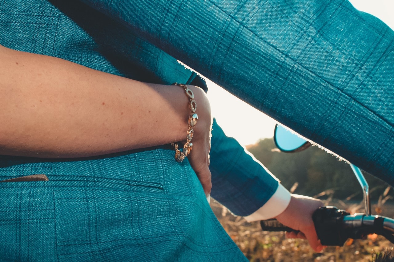 A couple's hands intertwined, with one partner wearing a gold bracelet, while the other is holding a bike handlebar, in front of a warm sunset.