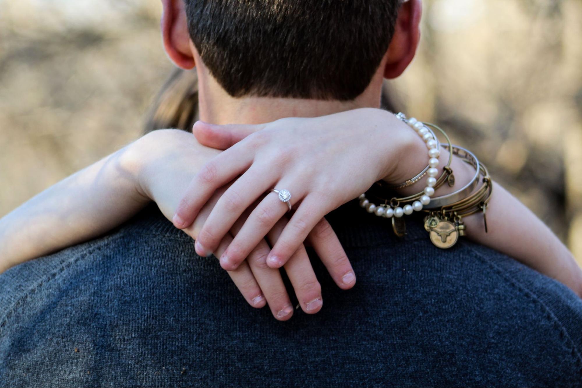 A woman hugs a man from behind, her hands resting on his shoulders and displaying a diamond engagement ring along with several bracelets, including a pearl bracelet and various bangles.