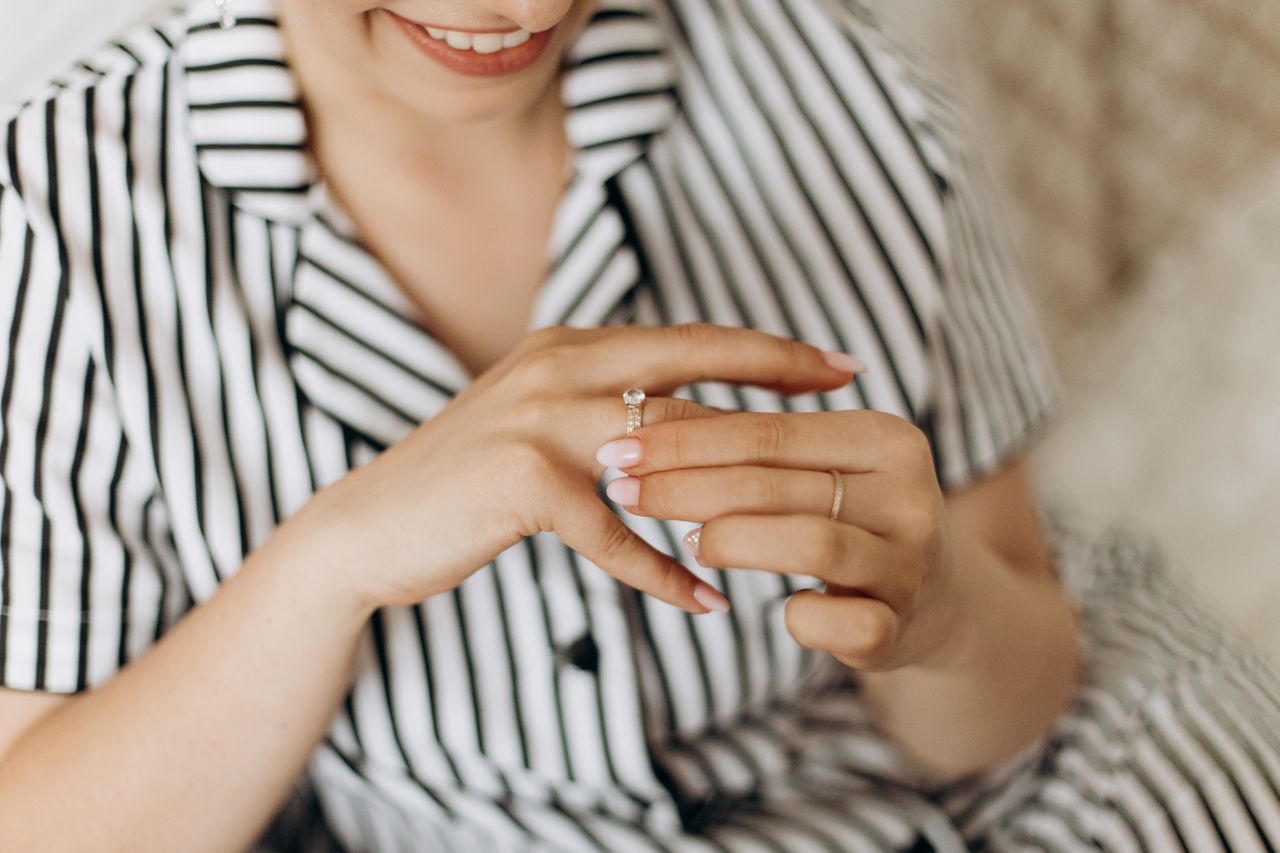 A close-up of a smiling woman in a striped shirt admiring a sparkling engagement ring on her finger.
