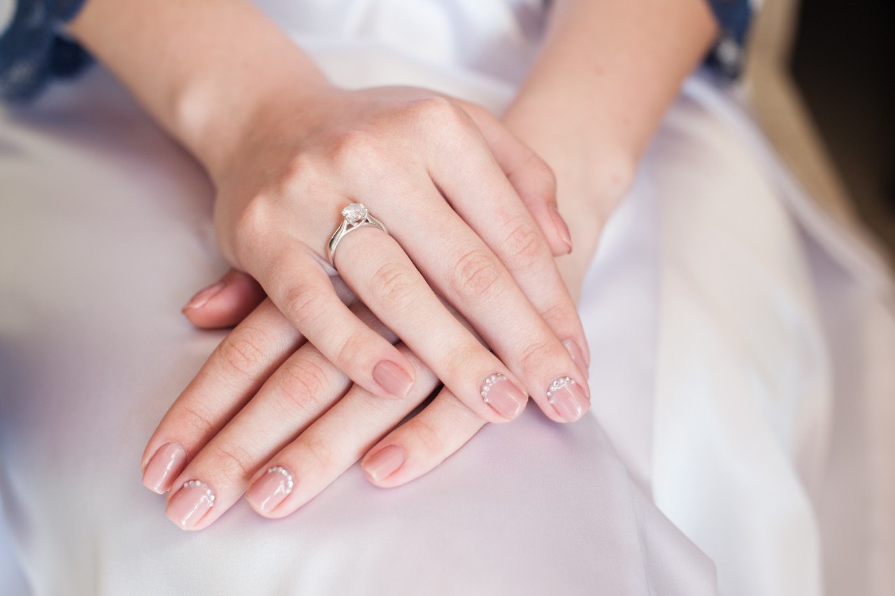 A close up of a bride's hands with delicate nail art and a diamond ring resting gently on a white gown.