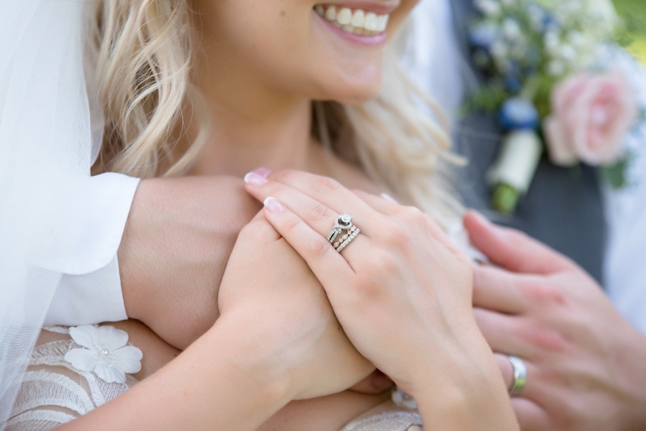 A close-up of a bride and groom holding hands, showing off their wedding rings. The bride is smiling, and there's a soft-focus floral bouquet in the background.
