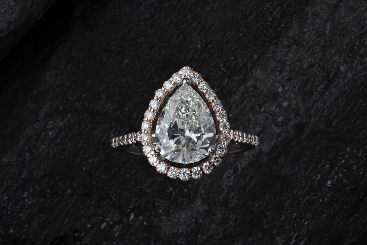 A pear-shaped diamond ring, surrounded by a halo of smaller diamonds, set against a textured black background