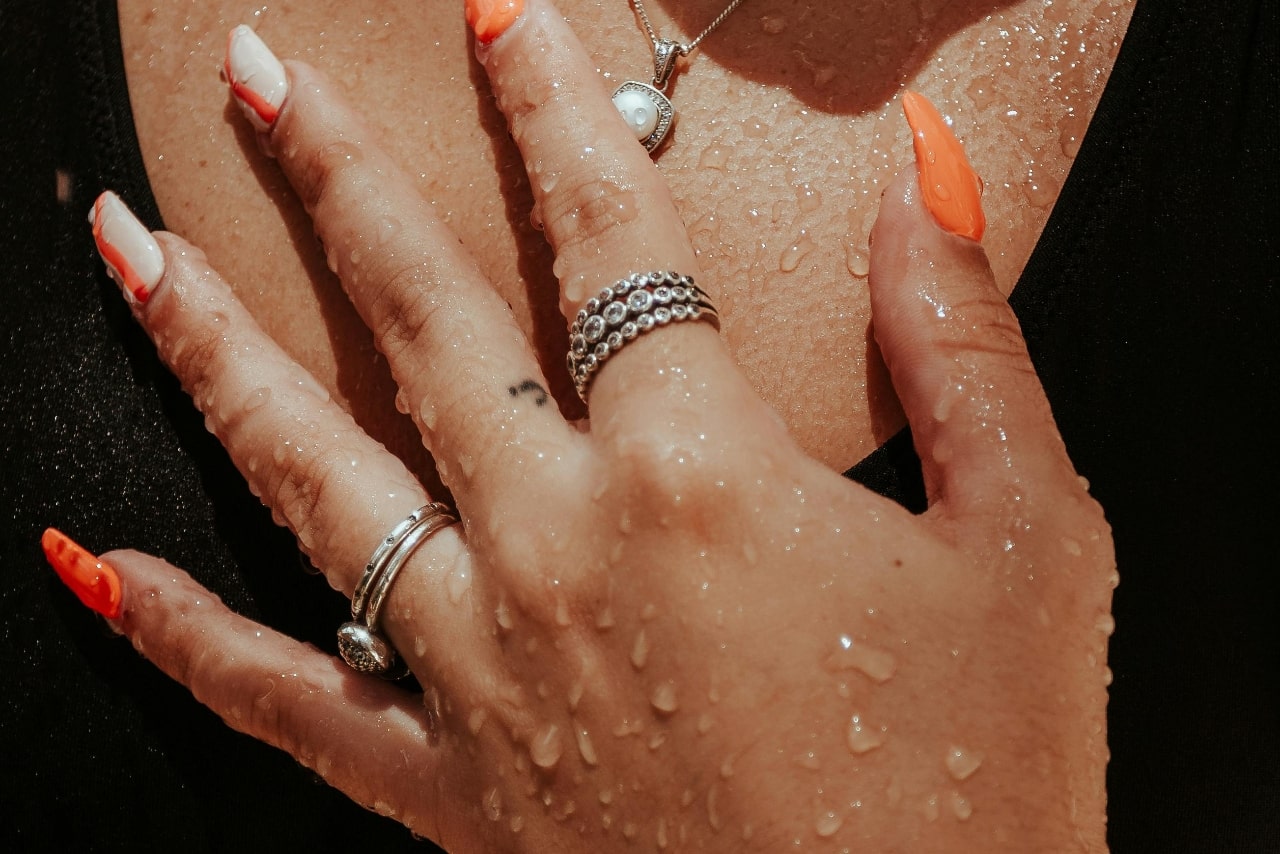 Close-up of a wet hand on a chest, showcasing orange and white manicured nails, layered silver rings, and a necklace. Close-up of a wet hand on a chest, showcasing orange and white manicured nails, layered silver rings, and a necklace.