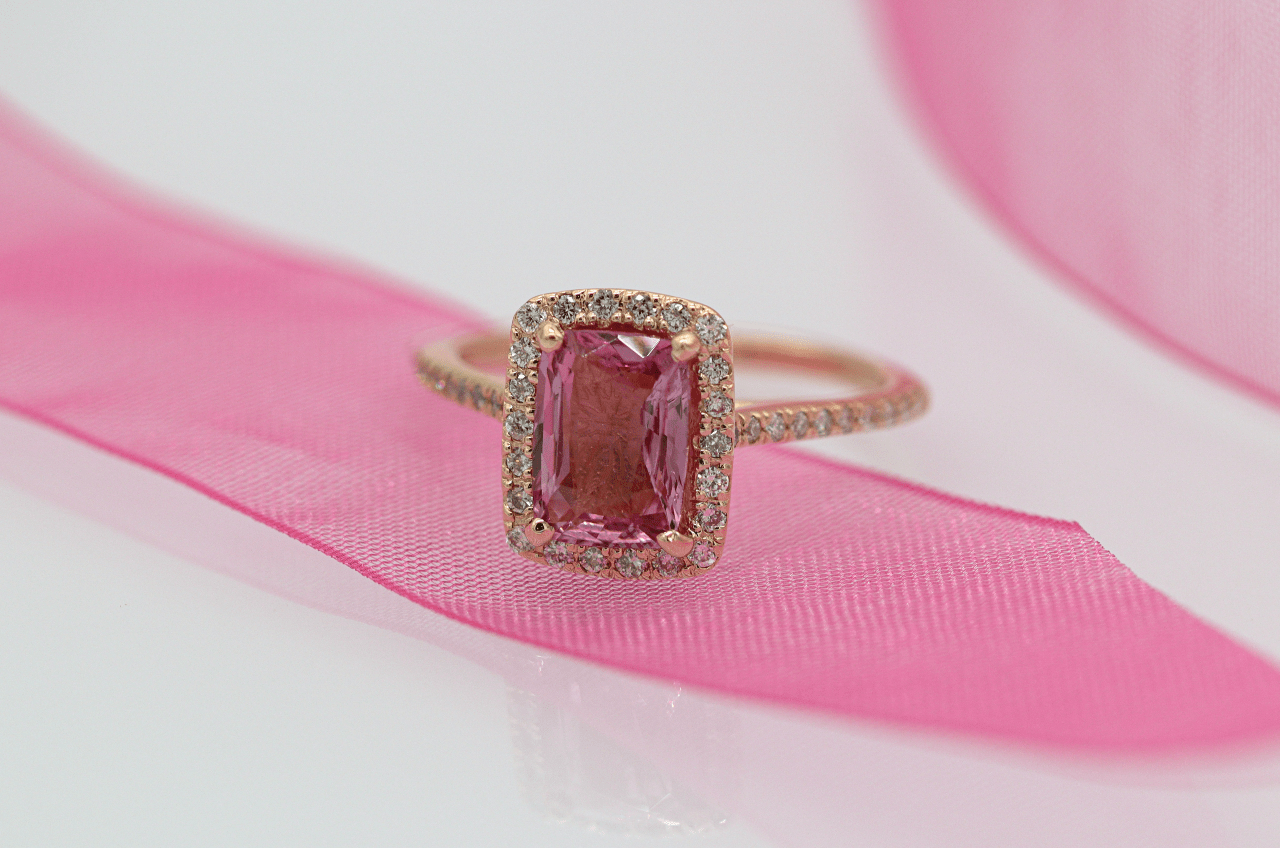 A rose gold ring with a radiant cut pink sapphire surrounded by small diamonds, rests on a pink ribbon. A rose gold ring with a radiant cut pink sapphire surrounded by small diamonds, rests on a pink ribbon.