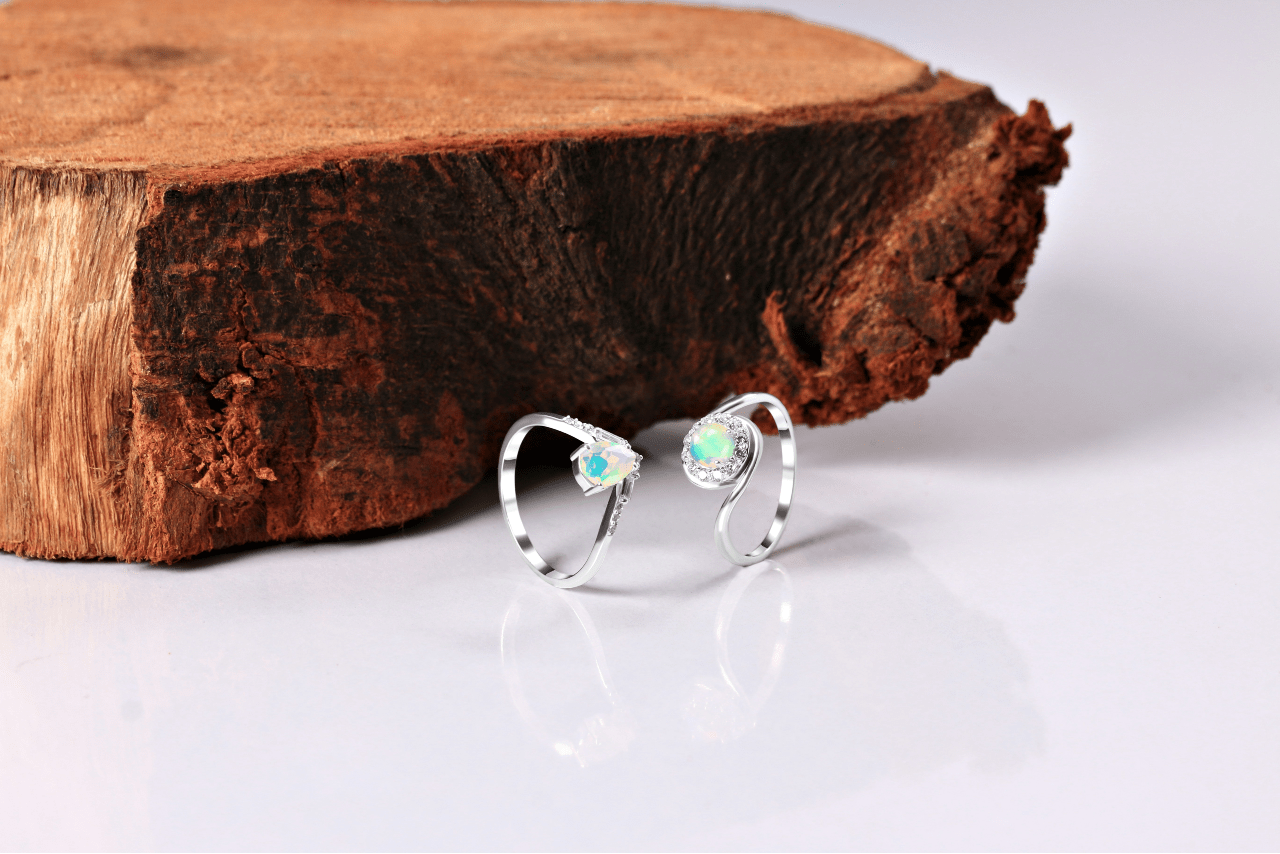 Two white gold freeform rings with opals sit on a reflective white surface with a rustic tree slab. Two white gold freeform rings with opals sit on a reflective white surface with a rustic tree slab.