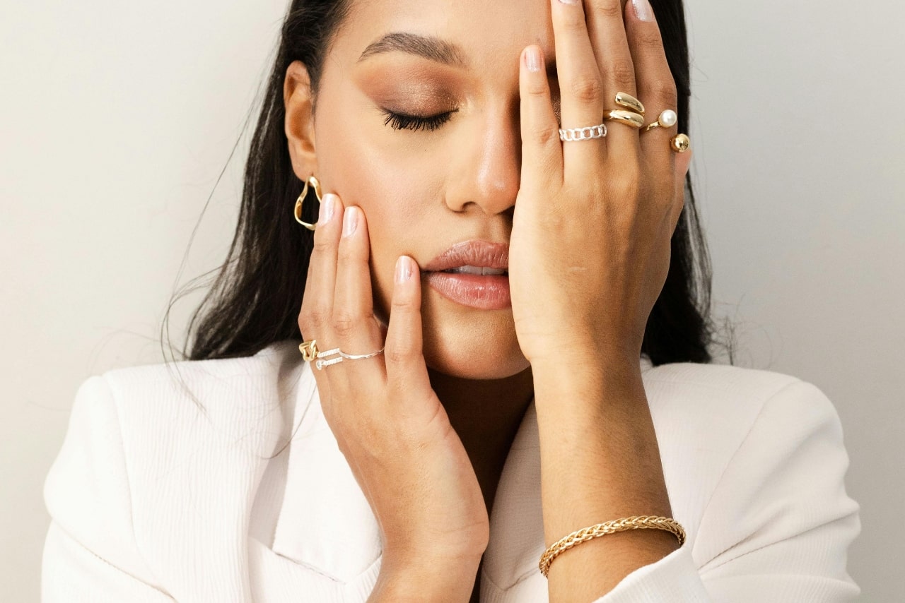 Woman in white blazer covers her face with one hand, displaying various yellow and white gold rings, a yellow gold bracelet, and contemporary gold hoop earring. Woman in white blazer covers her face with one hand, displaying various yellow and white gold rings, a yellow gold bracelet, and contemporary gold hoop earring.