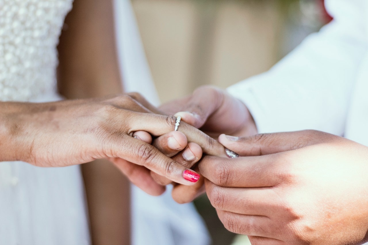 A man sliding a wedding band onto his bride’s finger, her nails manicured.