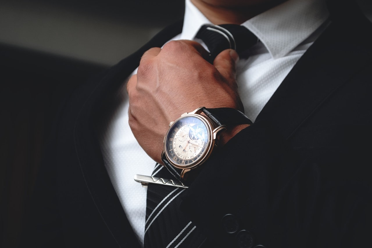 A man in a black suit fixing his tie, showcasing a black leather wristwatch with a moonphase complication. A man in a black suit fixing his tie, showcasing a black leather wristwatch with a moonphase complication.