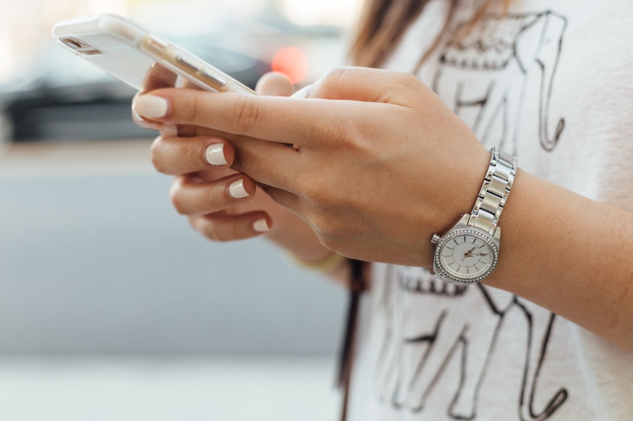 A woman with white manicure and t-shirt with elephant print typing on the phone, showcasing a stainless steel watch with white dial on her wrist. A woman with white manicure and t-shirt with elephant print typing on the phone, showcasing a stainless steel watch with white dial on her wrist.