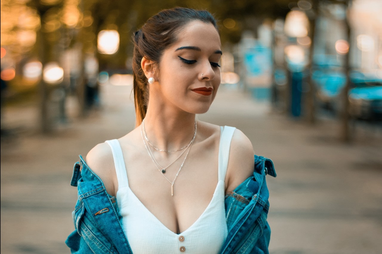 Stylish woman in a denim jacket adorned with layered necklaces and white stud earrings on a tree-lined street.