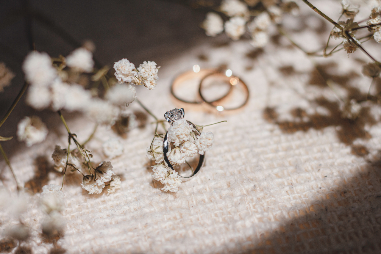 A platinum engagement ring lying on a cream-textured fabric surface with two yellow gold wedding bands in the background, surrounded by baby's breath.