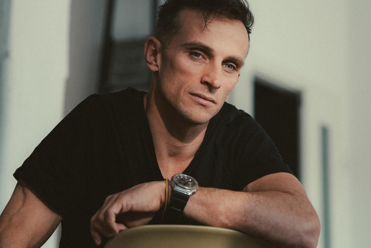A close-up of a casually dressed man in a black t-shirt wearing a sporty luxury watch. A close-up of a casually dressed man in a black t-shirt wearing a sporty luxury watch.