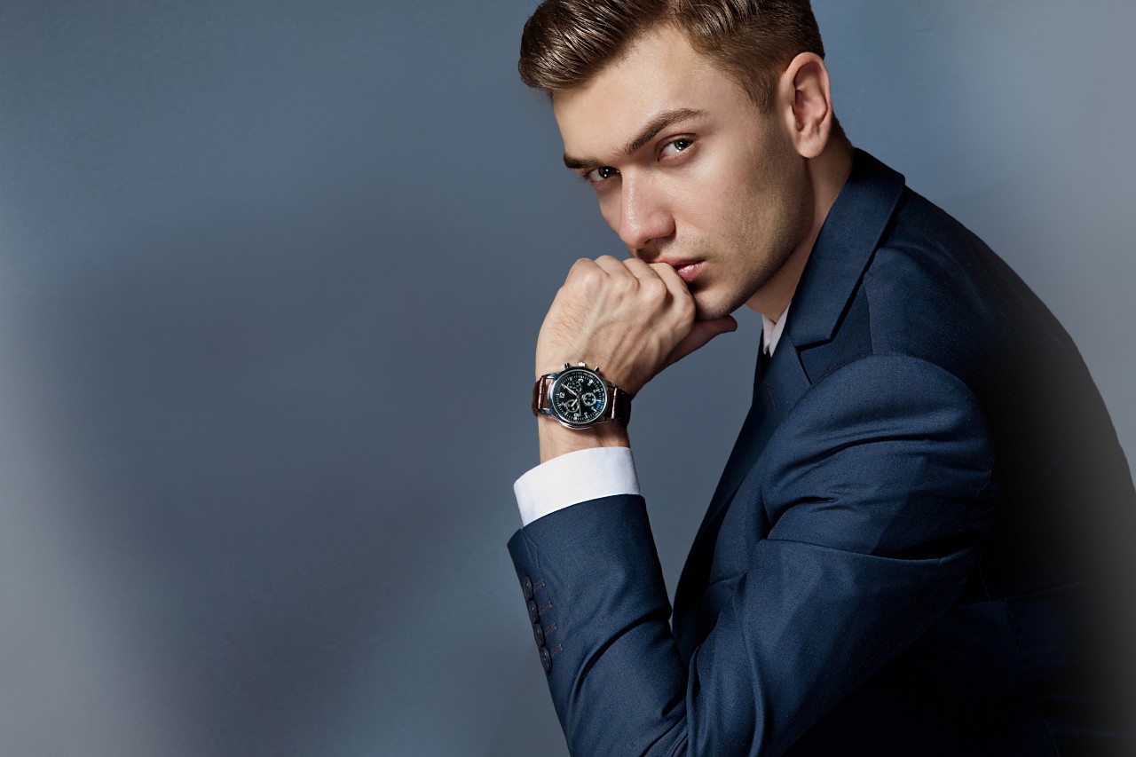 A man in dark grey suit puts his chin on his hand, showcasing brown leather wristwatch with black dial against a grey blurry background