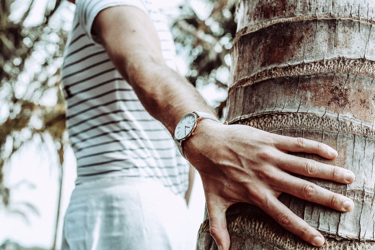 A close up of a man’s hand leaning on a tree, while demonstrating a minimalist wristwatch with white dial.