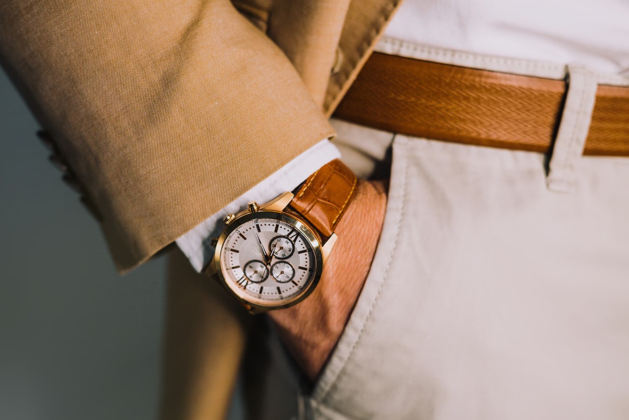 A man put his hand in a pocket of white trousers, demonstrating leather wristwatch with white dial and gold plated case.