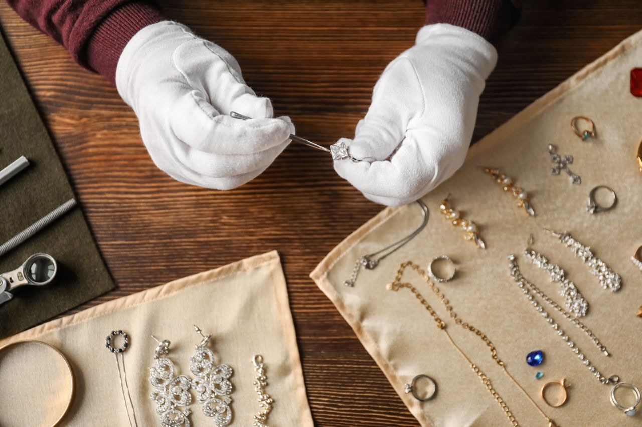 Close-up of a jeweler’s hands in white gloves carefully examining a diamond ring with tweezers, surrounded by jewelry arranged on beige cloths over a wooden table. Close-up of a jeweler’s hands in white gloves carefully examining a diamond ring with tweezers, surrounded by jewelry arranged on beige cloths over a wooden table.