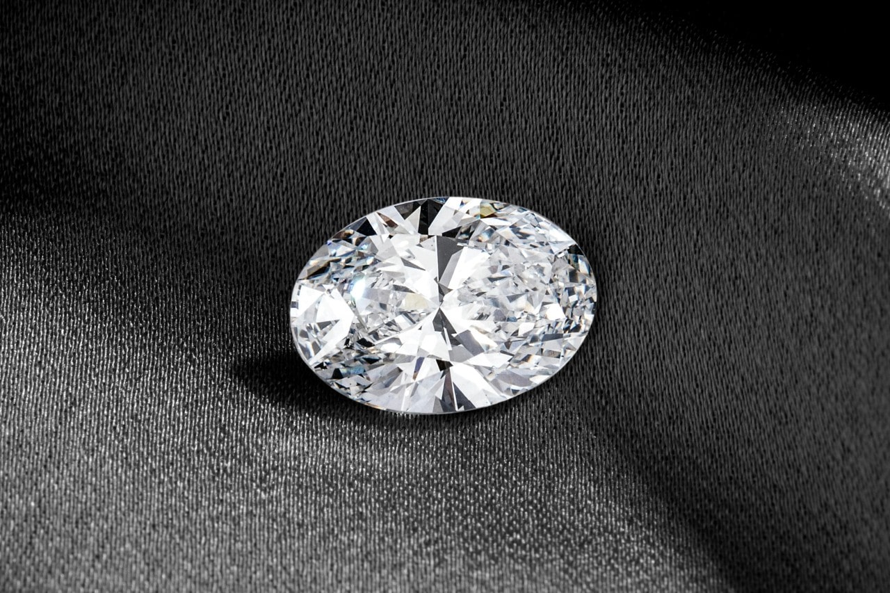 A close-up of a brilliant oval cut diamond shining while laying on a gray piece of fabric. A close-up of a brilliant oval cut diamond shining while laying on a gray piece of fabric.